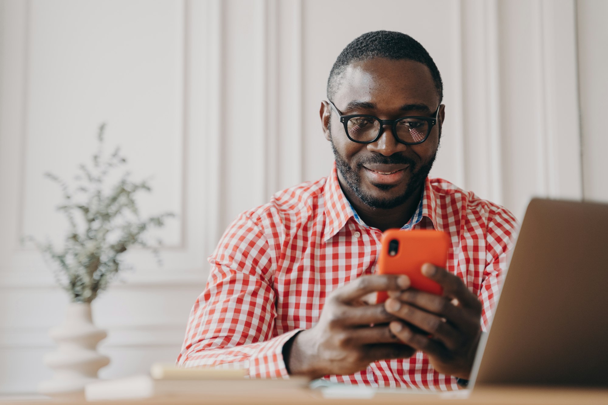 Smiling African ethnicity man office worker sitting at desk holding smartphone chatting with friends